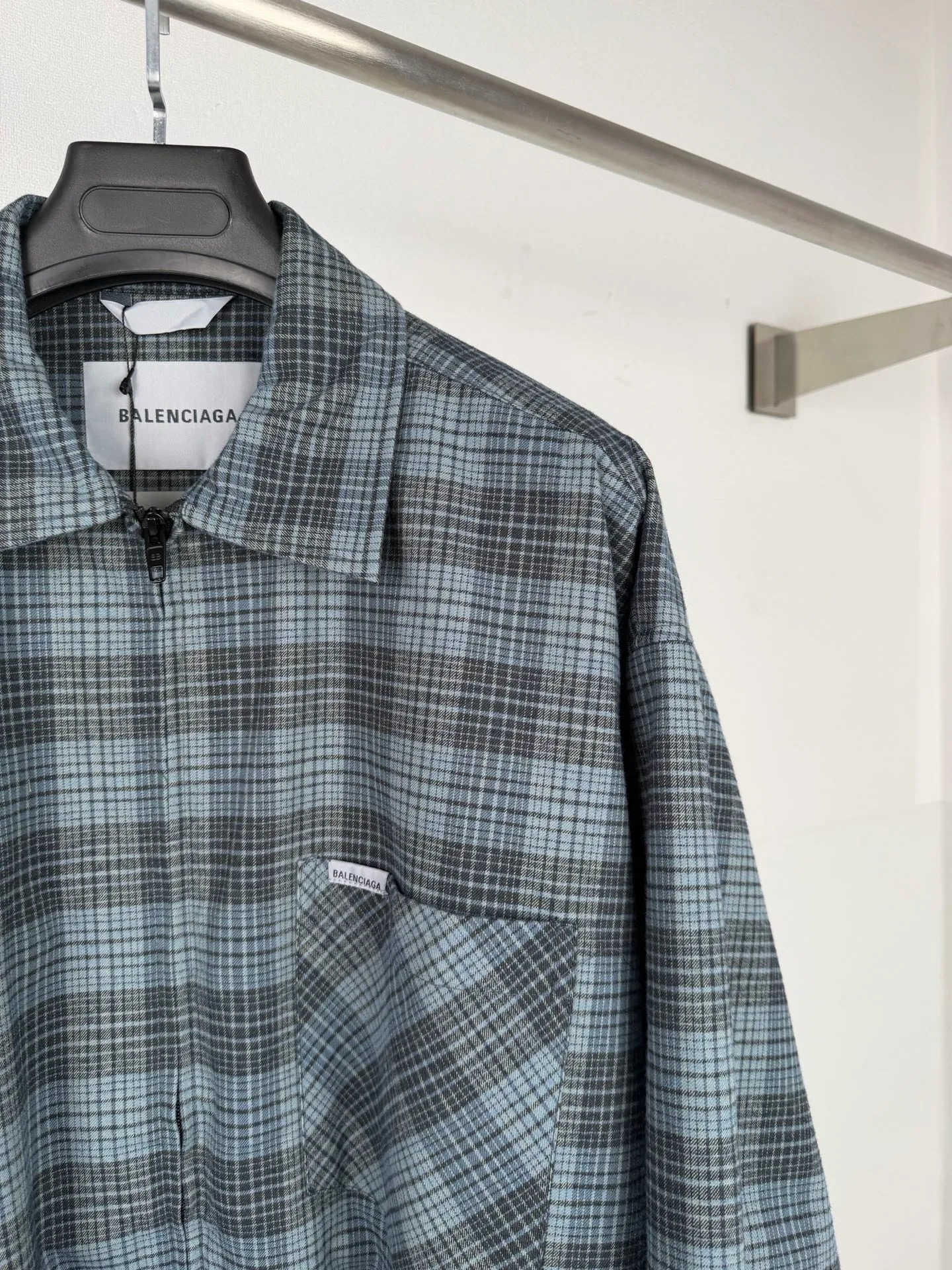 BALENCIAGA/balenciaga blue and gray plaid zipper pocket logo quilted jacket combines a three-layer structure of plaid woven fabric, filling cotton, and lining, and integrates plaid alignment technology and quilted garment production standards. There are multiple process difficulties in the whole process. The surface layer of the body is made of blue-gray Retro plaid fabric, and the cutting machine production strictly follows the global grid standards. The arrangement direction of the cutting pieces is fixed and cannot be changed at will. The horizontal and vertical lines of shoulder seams, side seams, and zipper placket joints must be consistent and coherent. The left chest patch pocket is cut independently, and the bag check pattern must be aligned with the front reference pattern. Slight deviations will destroy the visual integrity and directly increase fabric loss and cutting time. The most difficult part of the style is the three-layer composite sewing. The outer plaid fabric, filling cotton, and lining fabric have different physical properties, and the preshrinking processing parameters need to be adjusted separately. Pressure and temperature control are crucial during lamination operations. Once the parameters are out of balance, it can easily cause fabric stretch and distortion, plaid dislocation, and wrinkles and bulges in garments. At the same time, it is necessary to reasonably plan the quilting line to avoid the visual beauty of the grid pattern from being cut by the lines on the basis of firmly fixing the cotton layer and preventing cotton from running away. The placket adopts a centered zipper structure. It is difficult to sew the zipper tape and the quilted panels. It is required that the width of the left and right plackets be equal, the zipper routing is straight and smooth, and there is no lag in opening and closing. When the shirt lapel is filled with cotton, the thickness of the cotton needs to be controlled to prevent the collar from becoming bloated and unnaturally folded. The metal nameplate of the chest pocket is sewn at fixed points to avoid pulling the fabric during the operation and ensure that the plaid pattern is smooth and not deformed. The quilted material increases the overall thickness of the fabric. When sewing, you need to continuously adjust the presser foot pressure and stitch length to adapt to changes in sandwich thickness. The low-temperature and gentle ironing process is used for post-finishing to prevent the plaid from fading and the cotton layer from clumping due to high temperatures. From the precise alignment of cutting pieces, three-layer fabric pretreatment, composite quilting to garment finalization inspection, the processes are interlocking and the error tolerance rate is extremely low, testing the accuracy of plate making, equipment control and turning technology. Size:XS S M L